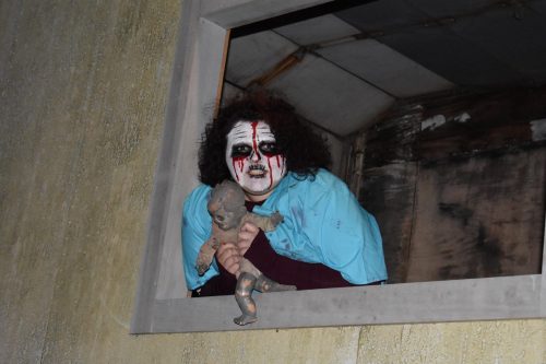 A person with spooky makeup and a doll peers out from a window, creating a chilling atmosphere.