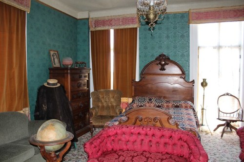 A vintage bedroom featuring a wooden bed, patterned textiles, a globe, and antique furniture against a teal wallpaper backdrop.