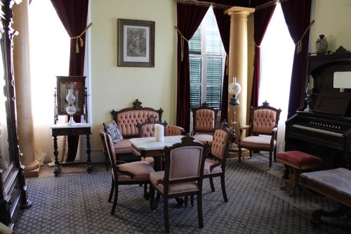 A vintage parlor with ornate furniture, a piano, and large windows draped with red curtains.