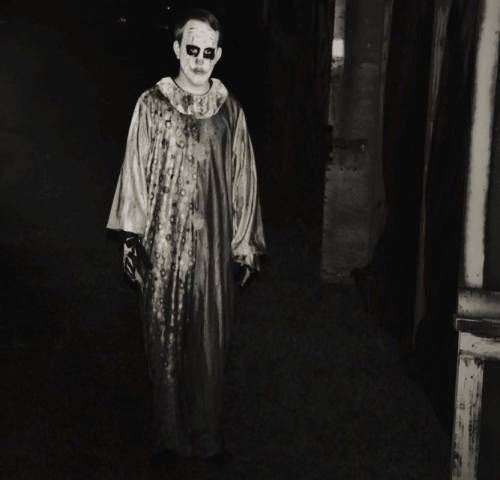 A creepy clown in a tattered costume stands in a dimly lit area, with a haunting expression and dark makeup.