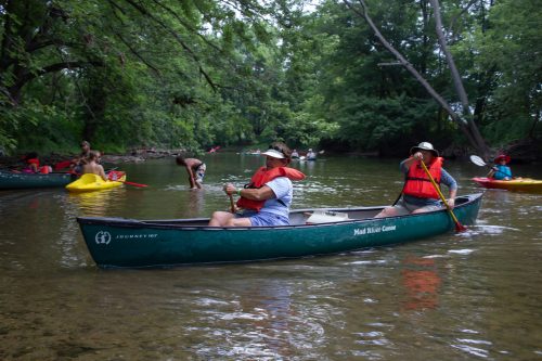 Two people in a canoe wearing life jackets paddle on a calm river, surrounded by trees and other kayakers.