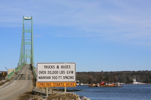 A green bridge spans a river, with a sign warning trucks and buses about weight limits and spacing requirements.