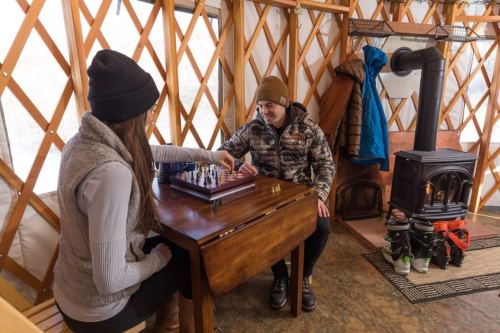 A man and woman play chess inside a cozy yurt, with a wood stove and winter gear nearby.