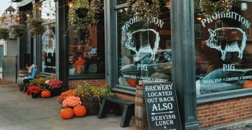 Exterior of Prohibition Pig brewery with pig-themed signage, pumpkins, and flowers, featuring a lunch menu sign.