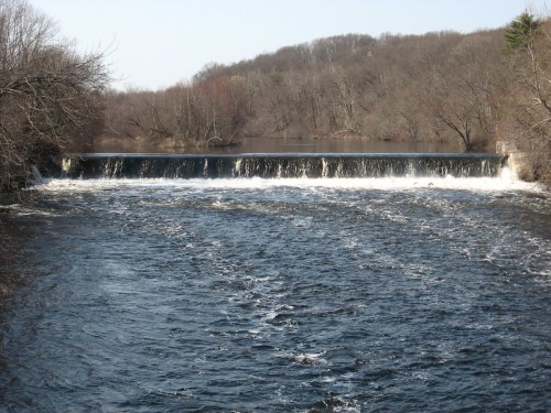 A serene river scene featuring a waterfall cascading over a dam, surrounded by bare trees and distant hills.