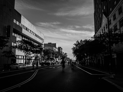 A black and white street scene with people walking, flanked by buildings and trees under a cloudy sky.