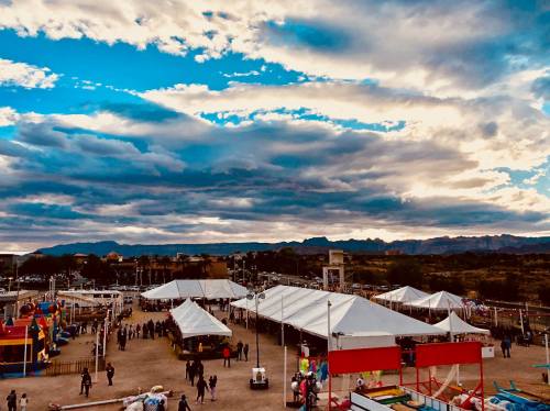 A bustling outdoor event with white tents under a dramatic sky, surrounded by mountains in the background.
