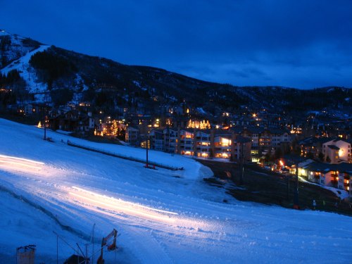 A snowy mountain landscape at dusk, with illuminated buildings and ski trails in the foreground.