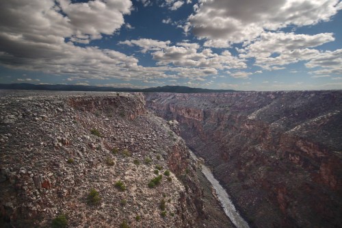 A vast canyon with rocky cliffs and a winding river below, under a sky filled with fluffy clouds.