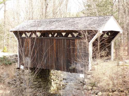 A rustic covered bridge made of wood spans a small stream, surrounded by bare trees and a peaceful landscape.