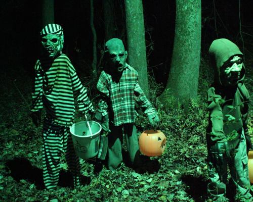 Three eerie, costumed figures in a dark, green-lit forest, holding candy buckets and a pumpkin.
