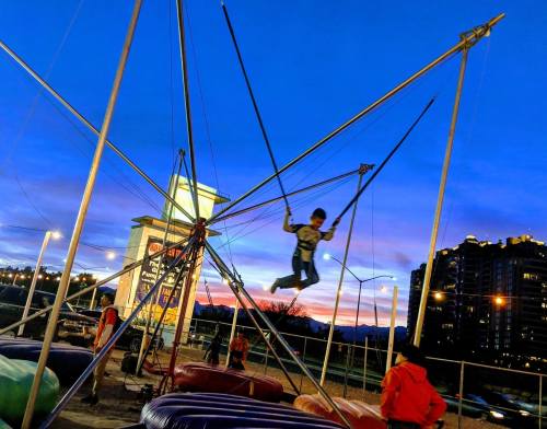 A child swings high on a bungee trampoline against a colorful sunset, with city buildings in the background.