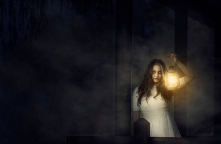 A woman in a white dress holds a lantern, standing in a dark, misty environment.