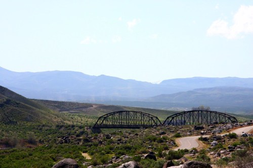 A black metal bridge spans a valley, surrounded by green hills and mountains under a clear blue sky.