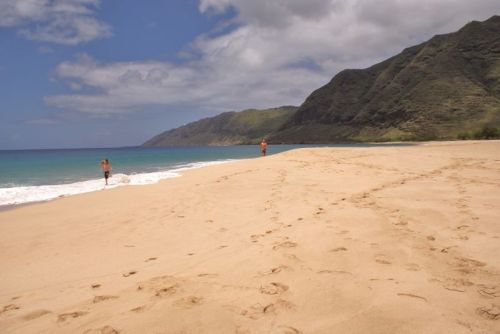 Two people walking along a sandy beach with gentle waves and a mountainous backdrop under a partly cloudy sky.