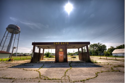 Abandoned car wash building with a water tower in the background, surrounded by overgrown grass and a cloudy sky.