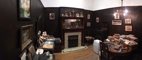 A vintage room with dark walls, a fireplace, framed photos, and stacks of old books and papers scattered around.