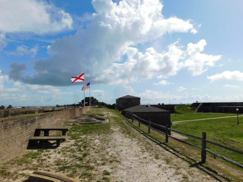 A scenic view of a historic fort with flags, grassy areas, and a blue sky filled with clouds.