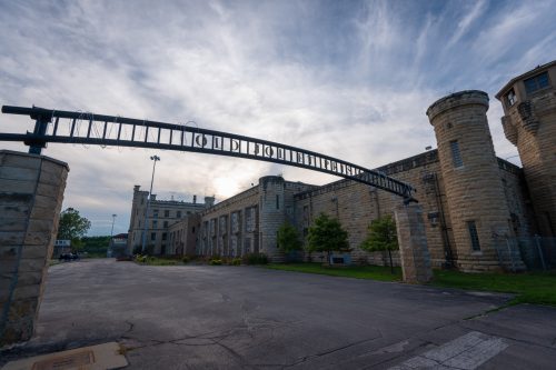 Entrance to an old stone prison with a barbed wire archway under a cloudy sky.