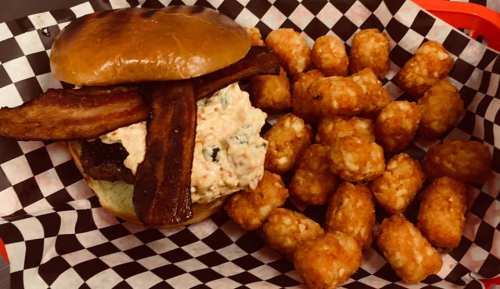 A burger with bacon and creamy topping, served with a side of crispy tater tots in a checkered basket.