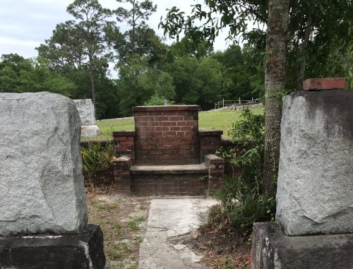 A bottle sits on a stone path between two large stone pillars, leading to a brick structure in a grassy area.