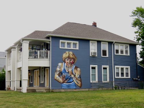 A blue house with a large mural of a woman with tattoos, holding a paintbrush, on its side. Grass in the foreground.