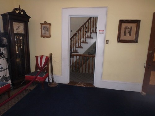 A hallway with a staircase, framed pictures on the walls, a vintage clock, and red chairs.