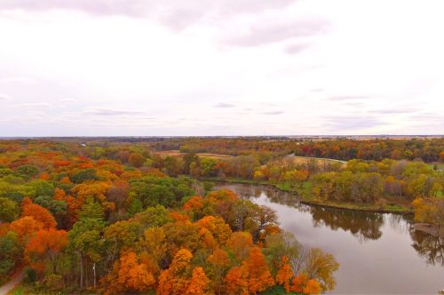 Aerial view of a river surrounded by vibrant autumn foliage in shades of orange, yellow, and green under a cloudy sky.