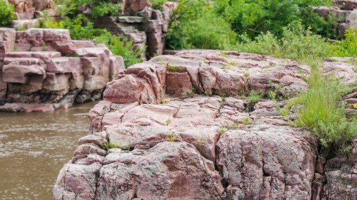 Rocky outcrop with greenery along a riverbank, featuring textured stone and lush vegetation in the background.