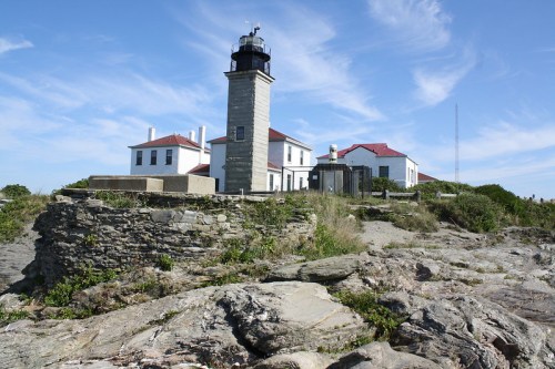 A lighthouse on a rocky shore, surrounded by greenery and a blue sky with wispy clouds.