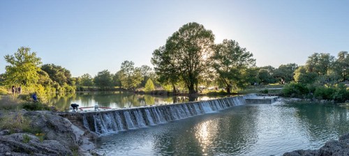 A serene landscape featuring a waterfall cascading into a calm river, surrounded by lush trees and a clear blue sky.