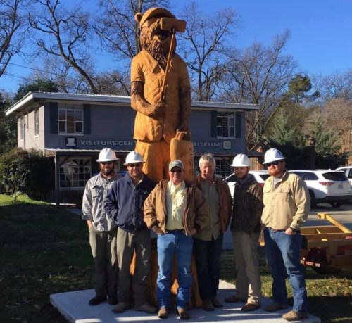A group of six men in hard hats stands beside a large wooden statue of a bear in front of a visitor center.