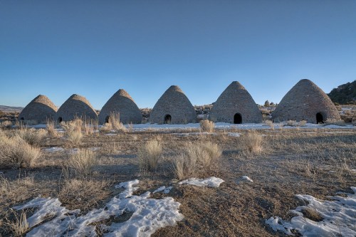 Six stone beehive-shaped kilns stand in a barren landscape, with sparse vegetation and a clear blue sky.