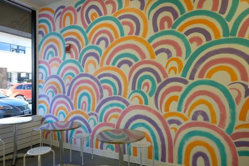 Colorful mural of overlapping rainbow arches in pastel shades on a wall, with small tables in front.