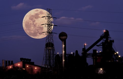 A large full moon rises behind industrial structures and power lines against a twilight sky.