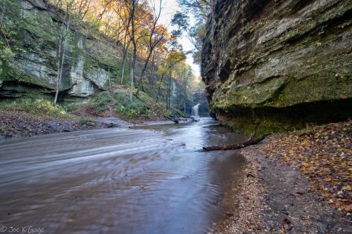 A serene river flows through a narrow canyon, with autumn foliage and a waterfall in the background.