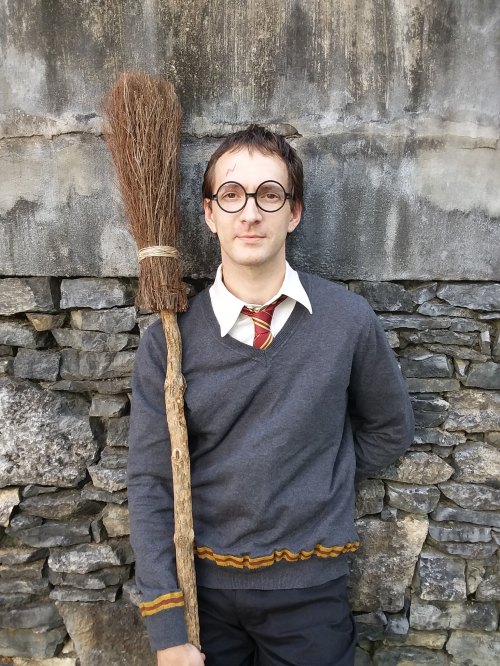 A person dressed as a wizard, wearing glasses and a gray sweater, holding a broomstick against a stone wall.