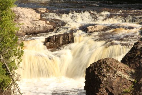 A cascading waterfall flows over rocky terrain, surrounded by lush greenery and a serene river setting.