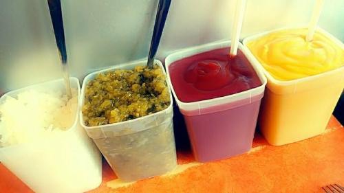 Four containers filled with different sauces: white rice, green relish, red sauce, and yellow sauce, with spoons and straws.