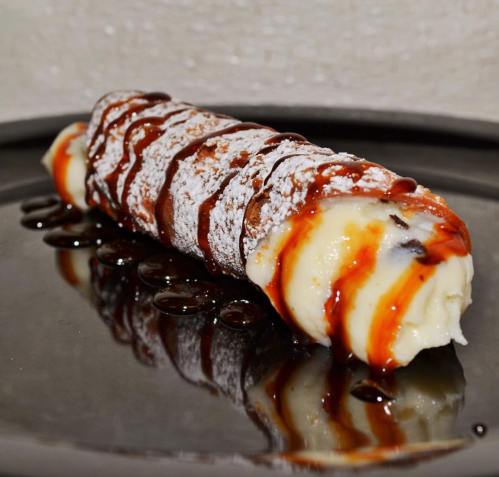 A dessert cannoli filled with cream, drizzled with chocolate sauce, and dusted with powdered sugar on a dark plate.