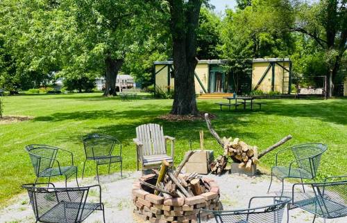 A fire pit surrounded by chairs in a grassy area, with trees and a building in the background.