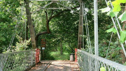 A metal suspension bridge surrounded by lush greenery, leading into a wooded area with a sign nearby.