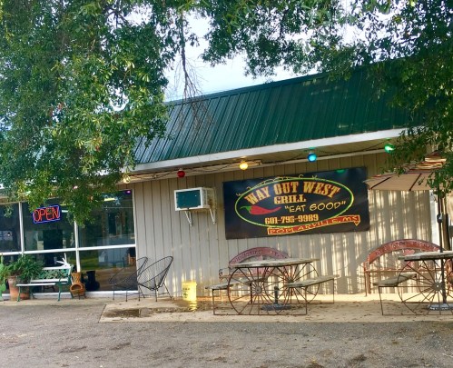 Exterior of a restaurant with a green roof, "Way Out West Grill" sign, and outdoor seating under trees.