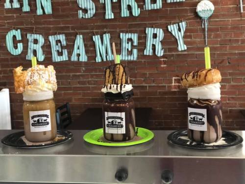 Three decadent milkshakes topped with whipped cream, cookies, and pastries, displayed on a counter with a brick wall backdrop.