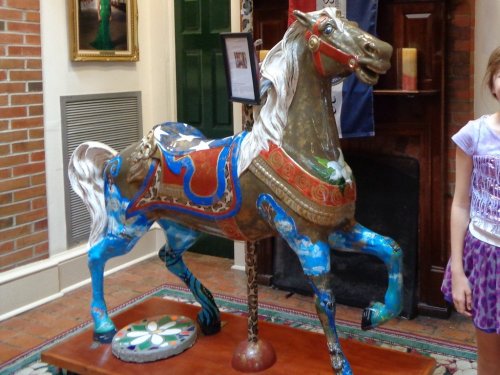 A colorful, ornate carousel horse displayed indoors next to a child, with a brick wall and fireplace in the background.