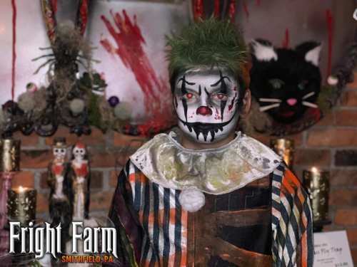 A clown with green hair and eerie makeup stands in front of a spooky backdrop, featuring candles and decorations.