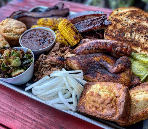 A platter of assorted barbecue meats, sides, and cornbread, featuring brisket, chicken, sausage, corn, and baked beans.