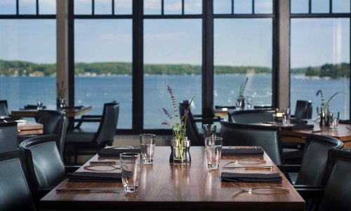 A dining area with wooden tables, black chairs, and a view of a lake through large windows. Fresh flowers on the tables.