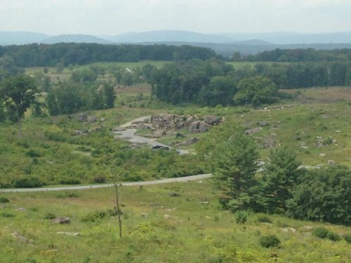 A scenic view of a grassy landscape with large rocks, trees, and distant mountains under a cloudy sky.