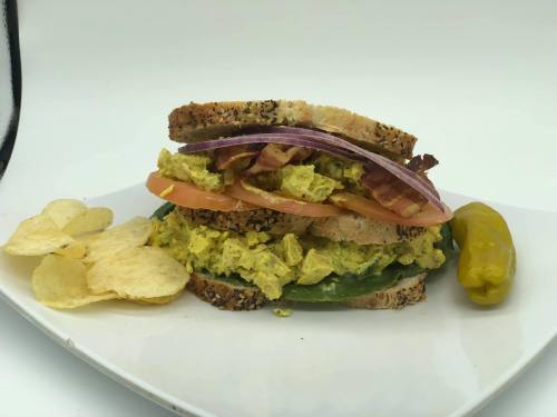 A stacked sandwich with egg salad, bacon, tomato, and onion, served with chips and a pepper on a white plate.
